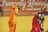 Dwie postacie stojące na tle baneru z napisem Koncerty Letnie. Jedna postać ubrana w kostium psa,…