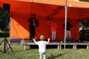 Na pierwszym planie małe dziecko z uniesionymi rękami, na dalszym planie zespół składający się z…
