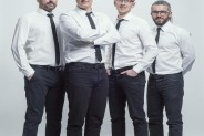 Na jasnym tle stoją czterej członkowie zespołu Albo i Nie. Mają czarne spodnie, białe koszule i…