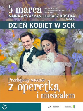 Na  pierwszym planie śpiewaczka i śpiewak w strojach wieczorowych. W tle wiosenne kwiaty. Poniżej…