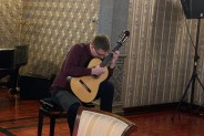 Młody mężczyzna w fioletowej koszuli w pozycji siedzącej, lekko pochylony nad klasyczną gitarą