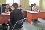 Za biurkami siedzą członkowie komisji kwalifikacyjnej. Po lewej stronie przed biurkiem, siedzący…