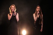 Na czarnym tle sceny dwie wokalistki Alona Szostak i Sofiia Popova śpiewają do trzymanych w…