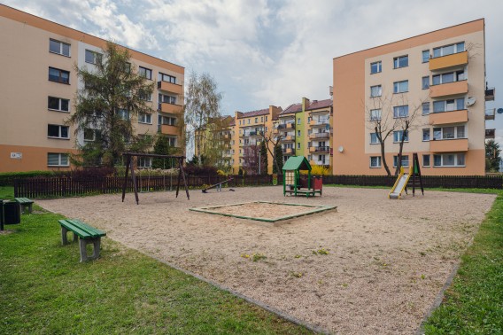 Osiedlowy plac zabaw z urządzeniami zabawowymi otoczony drewnianym ogrodzeniem.