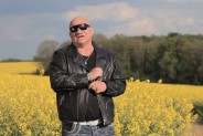 Na zdjęciu stojący na polu rzepaku piosenkarz Piotr Herdzina. Artysta ubrany jest na czarno, na…