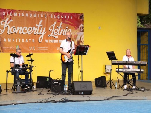 Na scenie amfiteatru, na tle żółtej ściany i czerwonego baneru stoją trzej muzycy, grają na…