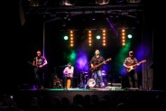 Na tle kolorowych świateł scenicznych zespół Macieja Balcara gra na swoich instrumentach a Maciej…
