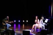 Po lewej stronie siedzi gitarzysta Adam Bul i gra. Po prawej siedzą dwie wokalistki. Jedna gra na…