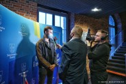 Michał Gabor pozujący na ściance z logotypami miasta. Przed nim stoi kamerzysta oraz osoba…