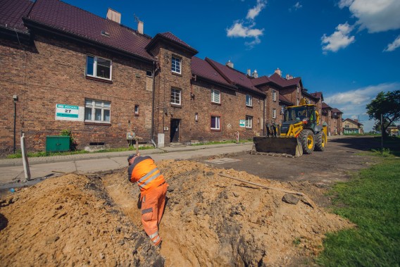Drogowiec wykonujący prace drogowe. Na drugim planie koparka.