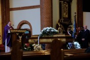 Po lewej ksiądz celebrujący uroczystości, w centrum trumna z ciałem zmarłego, po prawej w tle…