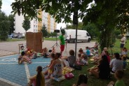 Grupka dzieci siedząca na trawie, w tle wiklnowy parawan, przed nim dzieci w kolorowych…