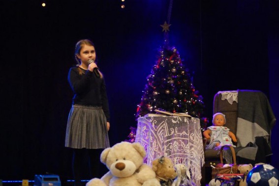 Wokalistka stoi na scenie trzymając mikrofon z ręce. ma czarną bluzkę i szarą spodniczkę. Za nią…