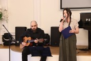 Mężczyna ubrany na czarno gra na gitarze obok dziewczyna z warkoczem śpiewa do mikrofonu