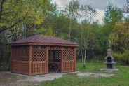 Drewniana altana wewnątrz, której stoi drewniany stół. Po prawej stronie znajduje się kamienny…