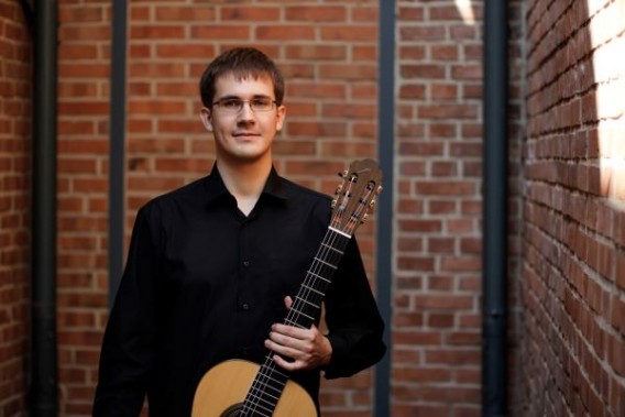 Na zdjęciu gitarzysta Wojciech Rysiecki stoi z gitarą, w czarnej koszuli, na tle ceglanej ściany.