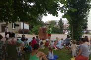 Grupka dzieci siedzących tyłem na trawie, patrzących na aktora ubranego w zieloną…