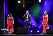 Tomasz Calicki śpiewa na scenie, trzymając w ręce mikrofon. Po obu jego stronach stoją dziewczyny…