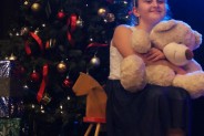 Wokalistka siedzi na scenie na bujanym koniku, trzymając w rękach misia. Za nią choinka.