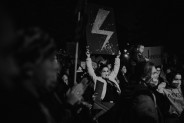 Kobieta w środku tłumu protestujących trzymająca baner z namalowaną błyskawicą, która jest…