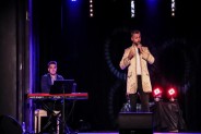 Na scenie Tomasz Białek ubrany w jasną marynarkę śpiewa do mirofonu. Obok niego siedzi pianista i…