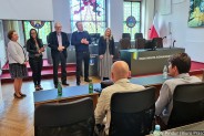 Młodzi sportowcy wraz z trenerami siedzą w dużej sali ratusza. Na drugim planie gospodarze…