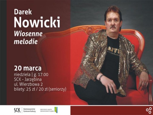 Na ciemnym tle czerwona kanapa. Na niej siedzi wąsaty mężczyzna w złotej marynarce i czarnej…