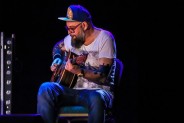 Na scenie gitarzysta zespołu Mikromusic siedzi i gra na gitarze. Ma białą koszulkę, brodę i…
