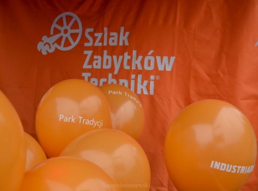 Na zdjęciu na tle pomarańczowego materiału z napisem Szlak Zabytków Techniki widzimy pomrańczowe…