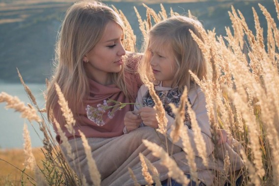 Mama i córka, obie jasnowłose siedzą wśród kłosów zbóż. W tle widać jezioro