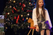 Wokalistka siedzi na scenie, na bujanym koniku. Za nią choinka.