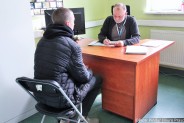 Członek komisji kwalifikacyjnej siedzący za biurkiem. Z przodu siedzący tyłem kandydat do służby…