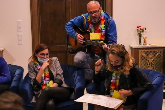 Mężczyzna w niebieskiej kurtce gra na gitarze jedna dziewczyna słucha druga zapisuje coś na kartce