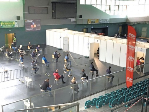 Ludzie siedzący na krzesłach ustawionych na płycie hali sportowej KS Michał oczekujący w kolejce…