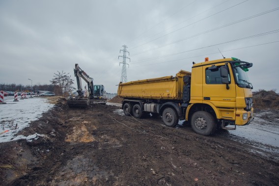Teren budowy łącznika drogowego. Na drodze stoi żółta ciężarówka i koparka.