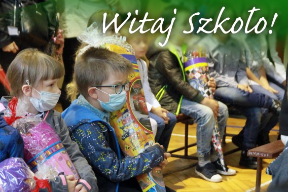 Dzieci z tytami siedzące w sali gimnastycznej, u góry napis "Witaj Szkoło!"