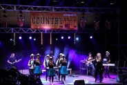Widok ogólny na scenę podczas Pikniku Country. Na scenie zespół tańca liniowego w białych…