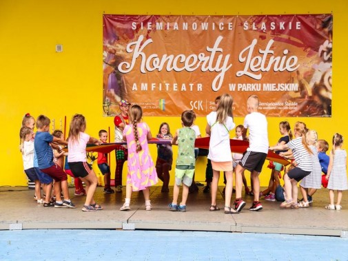 Dzieci bawią się na scenie amfiteatru podczas niedzielnych animacji. W tle żółta ściana…