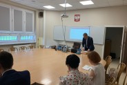 Wykładowca i słuchacze przy stole konferencyjnym, w tle tablica z materiałem pomocniczym,…