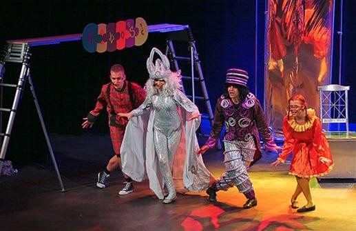 Czworo aktorów w bajkowych strojach tańczy na scenie teatru. W tle scenografia teatralna