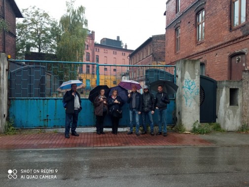 Grafika przedstawia grupę ludzi, stojących w deszczu z parasolami pod bramą dawnego browaru w…
