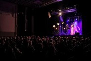 Widok na salę SCK-Bytków. Ludzie siedzą na widowni, na scenie kolorowe światła oraz dwoje…