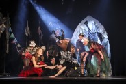 Grupa postaci w kostiumach nawiązujących do owadów w słupie jasnego światła na ciemnej scenie