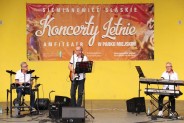Na scenie amfiteatru, na tle żółtej ściany i czerwonego baneru stoją trzej muzycy, grają na…
