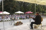 Widok na publiczność ze sceny. Widzowie siedzą na białych krzesłach i pod parasolami barowymi. W…