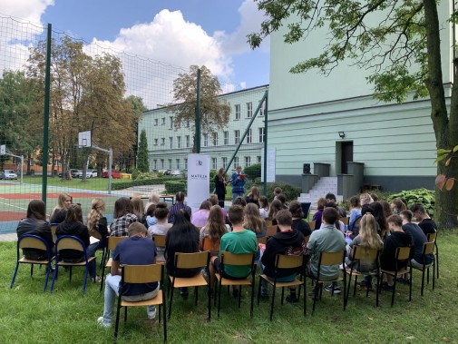 Uczniowie siedzący na krzesałach ustawionych na trawniku