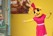 Gusia z rozłożonymi rękami. W tle scenografia teatru oraz żołta ściana, na której znajduje się…