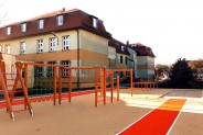 Urządzenia do kalisteniki oraz bieżnia do skoku w dal; w tle budynek szkoły