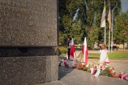 Dziewczynka w białej sukience pochyla głowę przed Pomnikiem Czynu Powstańczego.