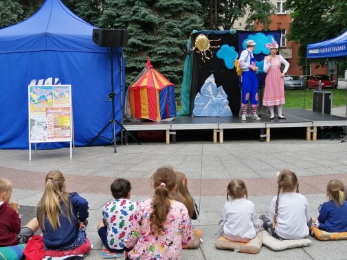 Dzieci siedzą na poduszkach na płycie rynku i oglądają spektakl. W tle scena z trójką aktorów,…